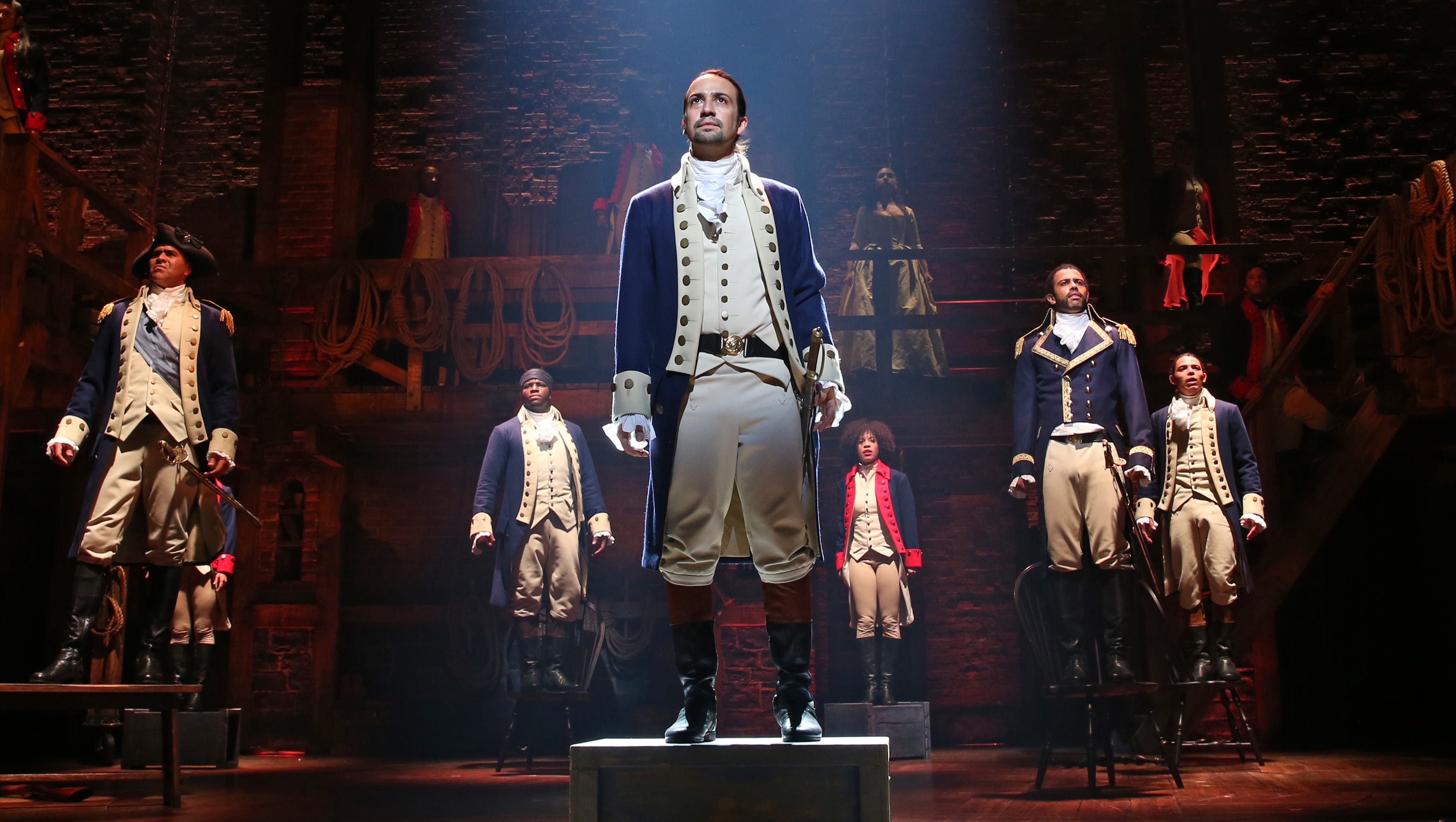 Image from the Broadway Musical Hamilton showing the title character in Revolutionary War-era costume flanked by other characters similarly dressed.
