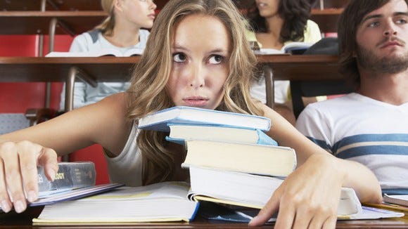 College student in lecture hall with chin on stack of textbooks, looking overwhelmed.