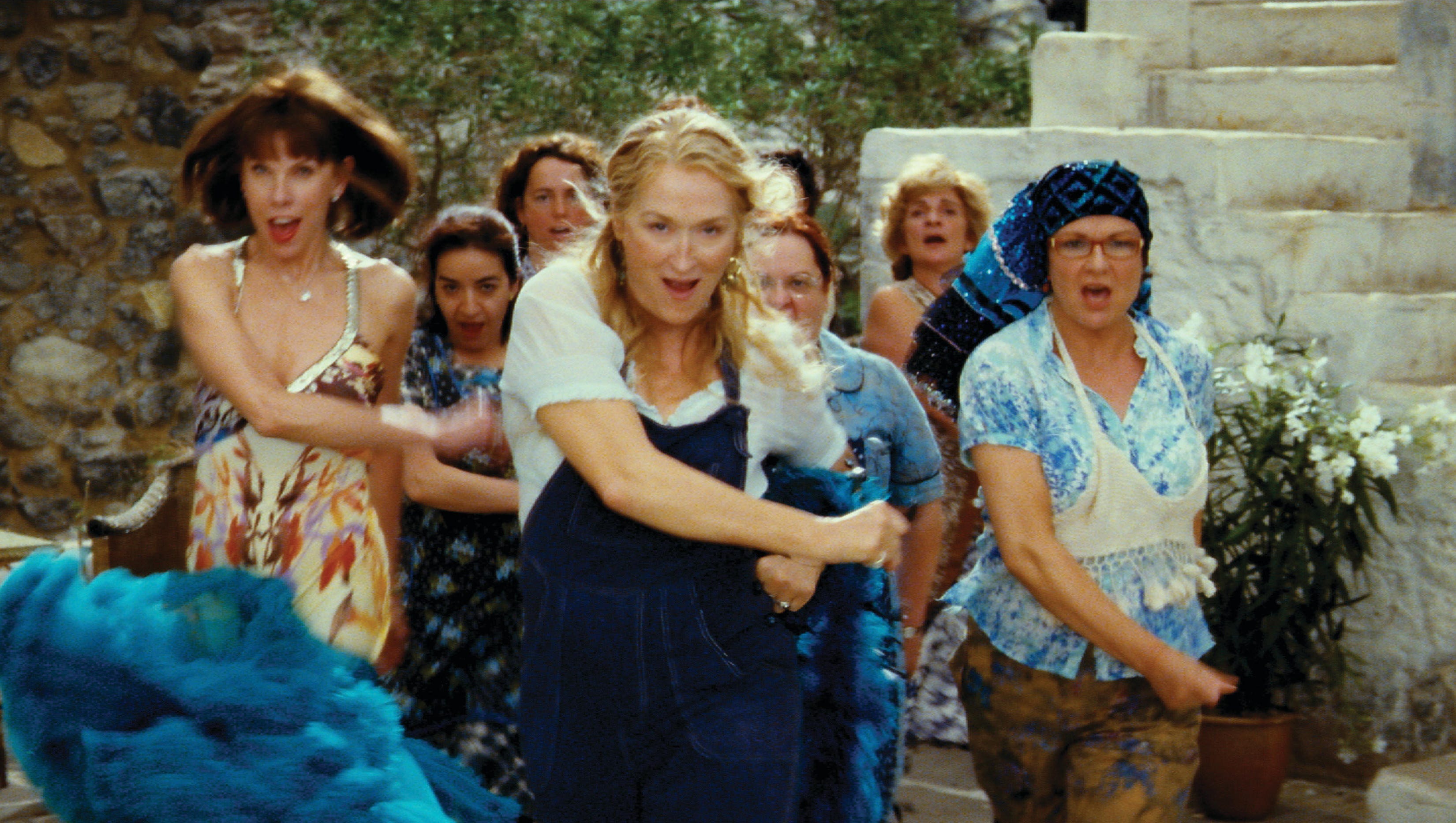 Image from the film Mamma Mia (2008). A cast of female characters confidently stride forward as they dance to ABBA's 'Dancing Queen against the backdrop of a Greek village.