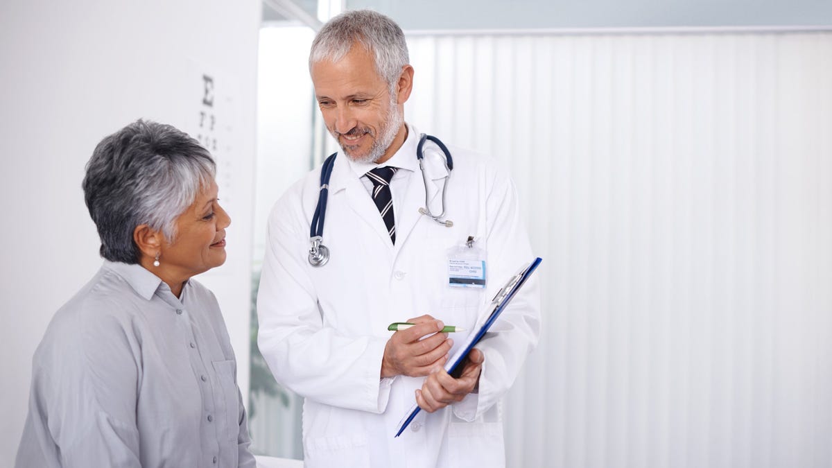 A person in a white coat and clipboards talks to a seated person.