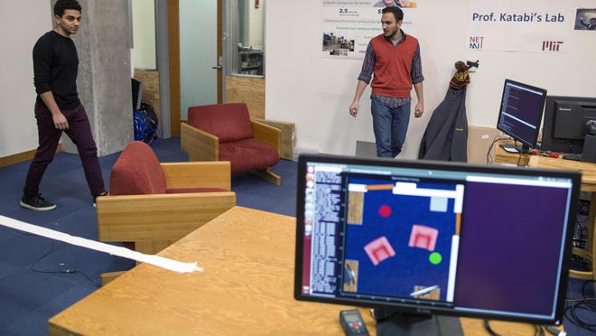 MIT researchers Emad Farag, left, and Fadel Adib walk through a room with chairs as a screen displays how RF-Capture is tracking their movements through the wall behind them, on the MIT campus in Cambridge, Mass.