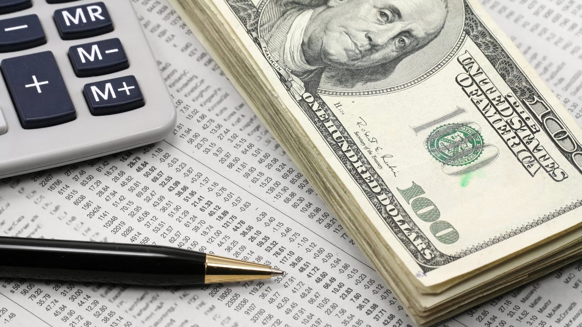 A neat stack of cash, a calculator, and a pen lying atop a financial newspaper with stock quotes.