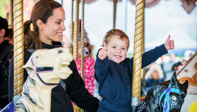 A happy mother and son are riding on a carousel together, smiling and having fun at an amusement park.  The boy holds two thumbs up.