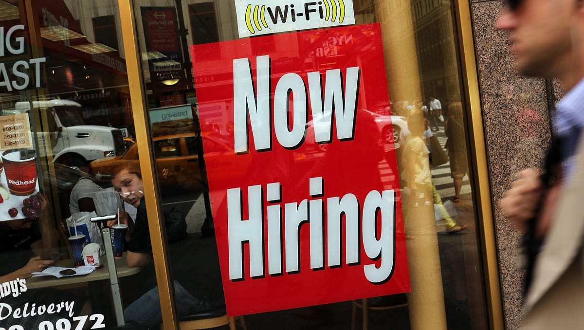 A "now hiring" sign is viewed in the window of a business.