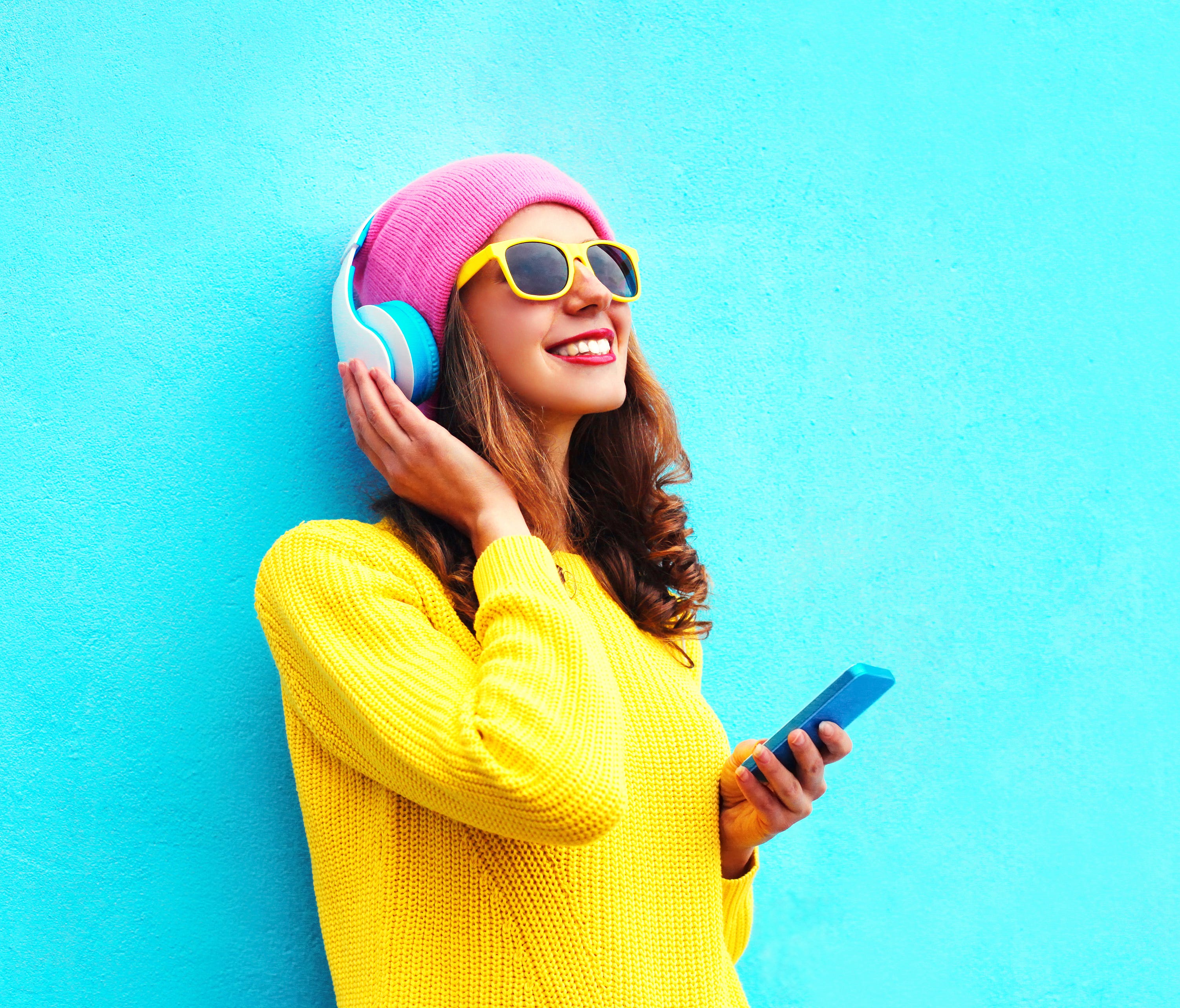 Fashion pretty sweet carefree girl listening to music in headphones with smartphone wearing a colorful pink hat yellow sunglasses sweater over blue background