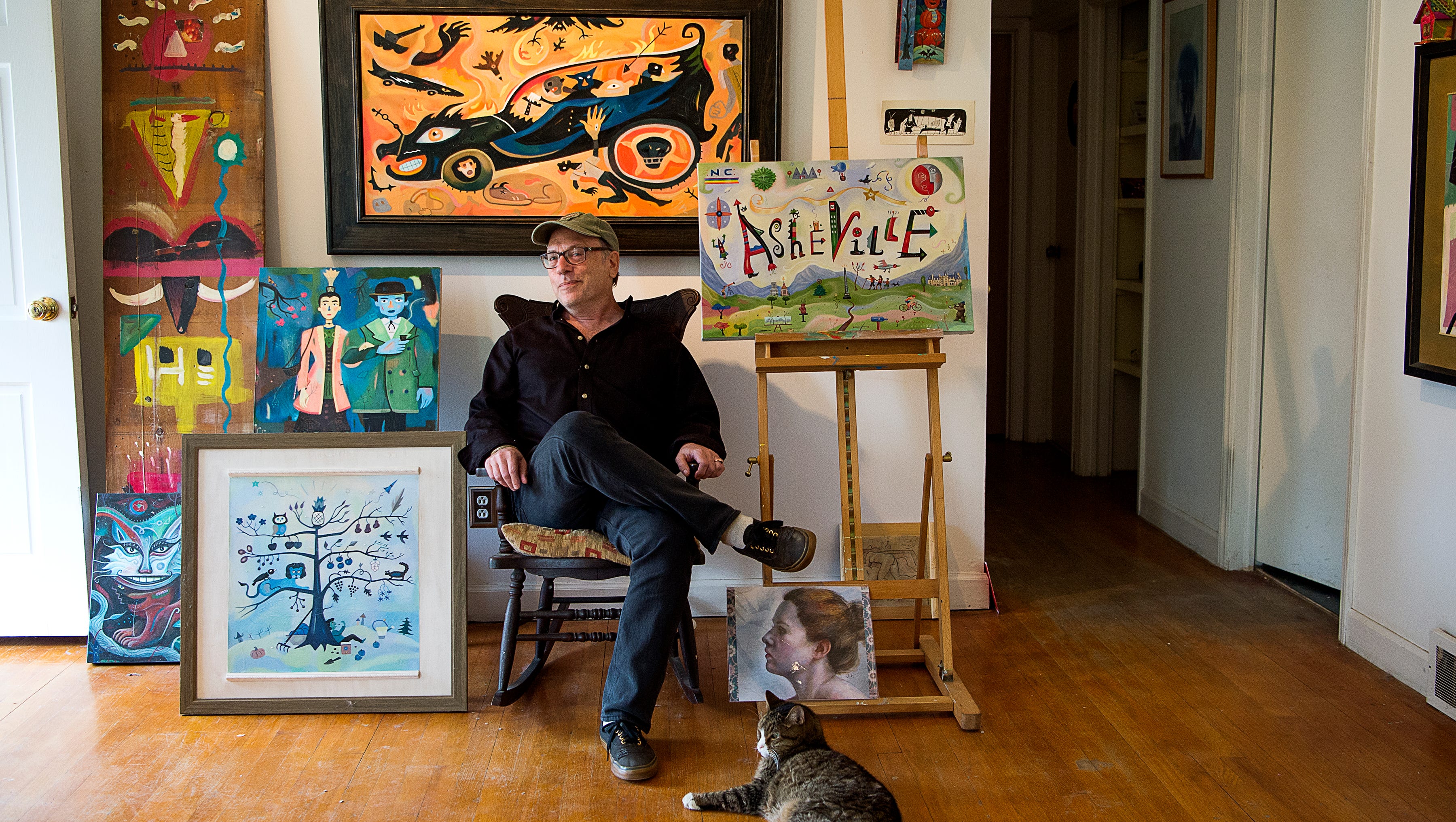 Andy Levine, an artist who works under the name John Nebraska, sits in his living room and studio surrounded by his artwork and cat in his Haw Creek home.