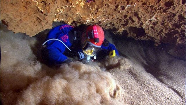 Exploration diver Tom Morris squeezes through a small crack in an underwater cave while sand blasts in his mask with the force of a fire hose.