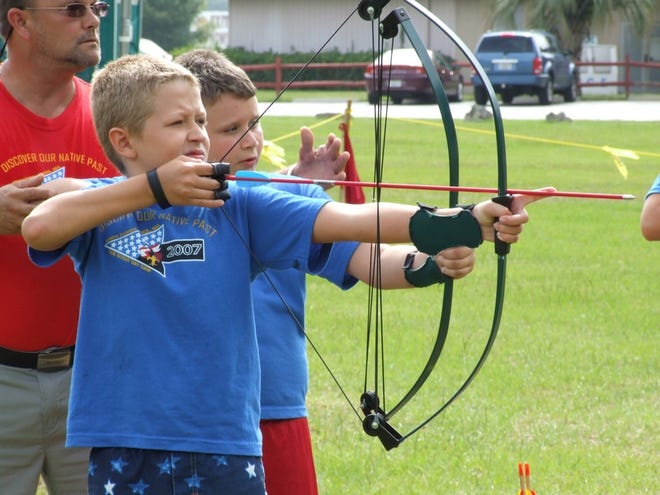 Oak Tree Village camp leader Dave Salcito helps Webelos Scout Joshua Hess, left, and Collin Mahan, right, at the Archery Range during the Cub Scout summer camp Wednesday. Below, Pack 197 Bear Cub Tristen Nappi carries his tribe's flag complete with their "doodles" of beadwork.
