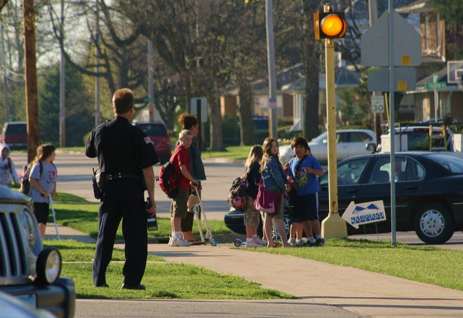 A Morton police officer holds a radar gun and watches Jefferson Street as students wait to cross the road on their way to school Friday. Morton police are stepping up speed enforcement near school zones as the weather transitions to warmer temperatures.