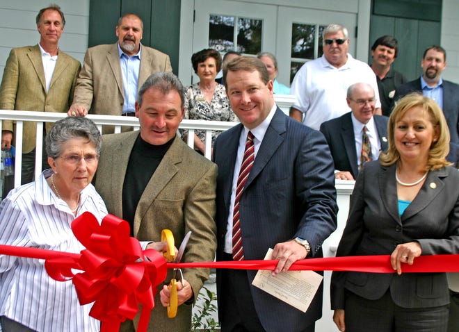 RIBBON CUTTING…Mrs. Monica Fleniken, left, is assisted by Iberville Parish Assessor Randy Sexton, second from left, as she cuts the ribbon to official open the new Iberville Parish Visitors Center located in Grosse Tete near I-10 last week. Finiken is a relative the original mayor of Grosse Tete, C.J. Finiken and the center is somewhat modeled after his residence. Looking on are Senator Rob Marionneaux and Representative Karen St. Germain. In the background on the front porch of the center are: Parish President J. Mitchell Ourso, second from left, and building contractor Edward Earl Comeaux and architect Bradley Guerin, far right, and other dignitaries.