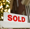 Real Estate "sold" sign with red brick building and trees blurry in the background