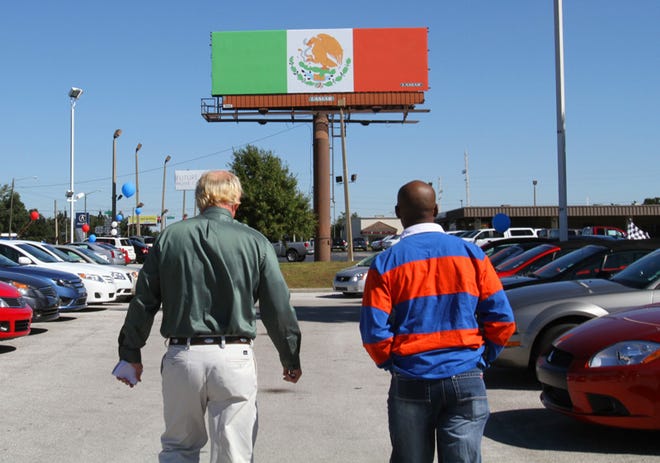 A salesman walks with a customer through the Ocala Nissan/Mitsubishi car lot on Southwest College Road, with a Mexican flag billboard in the background.
