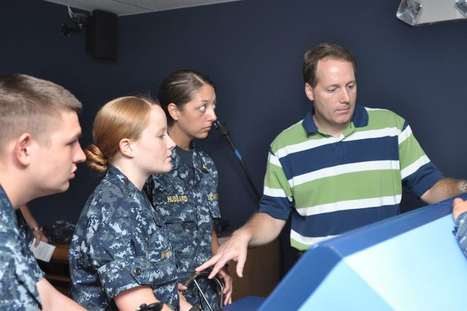 First Class Midshipman Charlotte Wygant, left, and First Class Midshipman Brittany Hubbbard, middle, listen closely to the instruction from BSH trainer operator John Carnley, right, during their turn working with the simulator.