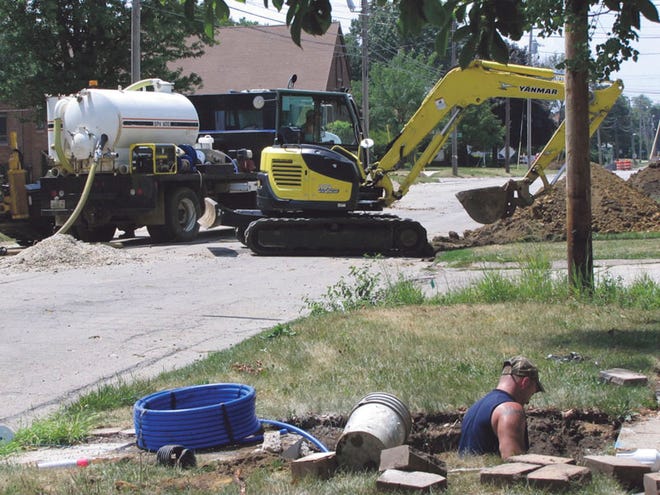 A contractor’s employee in the foreground hooks up the new water main to a home water service on East Division Street, while in the background an excavator works.