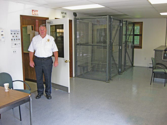 Dighton Police Chief Robert MacDonald stands in the entryway of the rear room of the Dighton Police Station that contains the two holding cells, the booking area (right) and the interview area (table to the left).