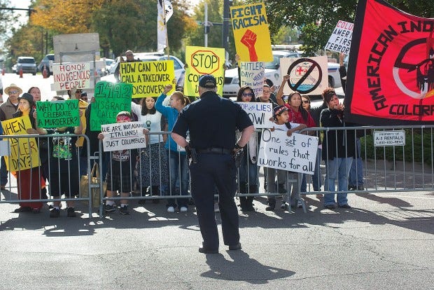 A Pueblo Police officer stands watch as protesters of the Columbus Day celebration holds signs as shout anti Columbus chants. Chieftain photo John Jaques 10.8.12