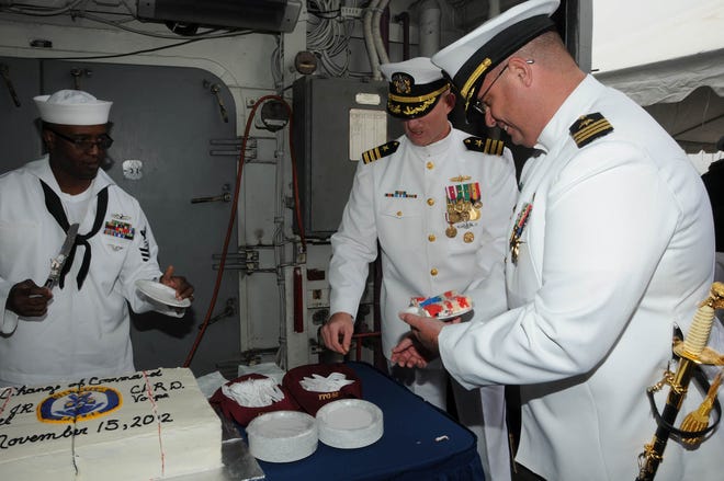 USS Taylor new Commanding Officer, Cmdr. Dennis Volpe, right, holds a piece of cake baked by the ship's culinary specialists to resemble the American flag when cut as outgoing Commanding Officer, Cmdr. Jeremy Hill, middle, reaches for a plate.