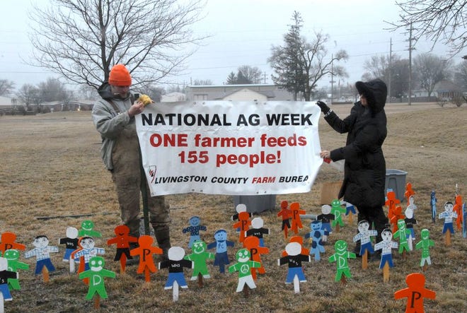 Dan Jones, a member of the Livingston County Farm Bureau’s Young Leaders, and Farm Bureau Manager Teresa Grant-Quick unfold a sign to remind the public about National Agriculture Week. They are standing among dozens of little wooden people placed to show people how many one farmer can feed. A close-up of the wooden people, painted by county FFA chapters, is below.