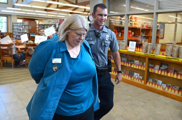 BRIAN D. SANDERFORD TIMES RECORD Van Buren police officer Brad Lipe escorts Van Buren Library director Danalene Porter out of the library as patrons protest after a mock arrest on Monday, Sept. 223, 2013. The library is conducting several events to bring attention to National Banned Book Week. Porter was reading The Wizard Of Oz to children prior to the "arrest." Porter said that book was banned in all Chicago public libraries in 1927. The library has a display, at right, of more than 60 books that have been banned at one time including The Great Gatsby, Catch-22, and recently the Harry Potter series.