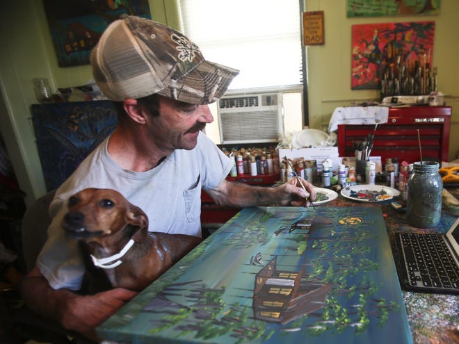 Lockport artist Hank Holland puts finishing touches on a painting at his home Friday while holding his paint-splattered pooch Sugar.