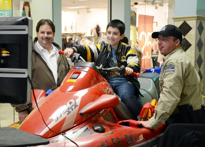 Issac Shipp, middle, rides a Jet Ski simulator while his grandfather, Michael Shipp, left, both of Nevada, Mo., and Kansas Department of Wildlife and Parks natural resource officer Jeremy Stenstrom, right, watch on during the 8th annual Albers Marine Hunting and Fishing Show at Meadowbrook Mall on Saturday. ANDREW NASH/THE MORNING SUN