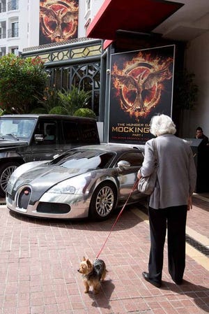 In this photo taken on Monday, May 12, 2014, a woman walking her dog looks at a Bugatti car and a poster for the “Hunger Games, Mockingjay: Part 1” at a hotel in Cannes, France.