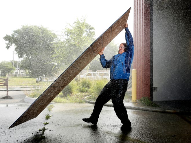 Jim Cantore is holding a 7/16" piece of OSB plywood, weighing about 45 to 50 pounds, in simulated 50 mph wind and rain to demonstrate the difficulties of boarding up for a hurricane too late.