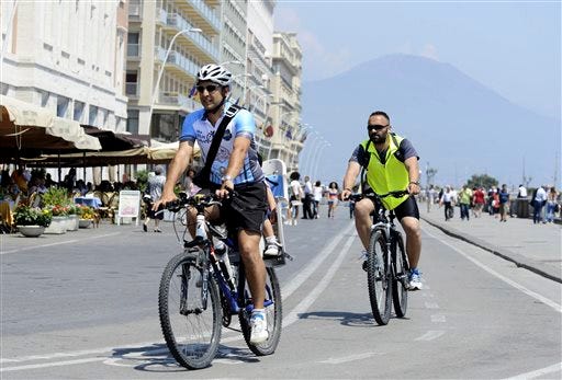 In this Friday, June 13, 2014 photo, Luca Simeone, founder of bike-sharing start up called "bike tour Napoli," left, pedals along a cyclists' path in Naples, Italy. In the background is Mt. Vesuvius. Three years ago Naples' seafront was an urban highway, noisy and smoggy, jammed with car traffic, while smelly trash erupted from garbage bins along streets and alleys. Urban cyclists were regarded as eco-fundamentalists. Three years later, Naples has a new mayor, clean streets, a wide pedestrian beachfront and a 20-kilometer (20-mile) cycling lane overlooking a beautiful bay. (AP Photo/Salvatore Laporta)