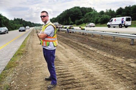 Levi Byers of the N.H. DOT waits for traffic to clear before he crosses Route 101 as the guardrail project expands into Exeter. The work is subcontracted out but DOT is overseeing it.