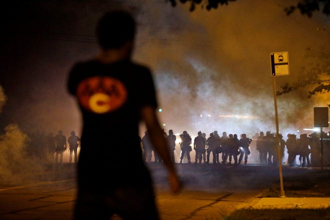 A man watches as police walk through a cloud of smoke during a clash with protesters Wednesday, Aug. 13, 2014, in Ferguson, Mo. Protests in the St. Louis suburb rocked by racial unrest since a white police officer shot an unarmed black teenager to death turned violent Wednesday night, with people lobbing Molotov cocktails at police who responded with smoke bombs and tear gas to disperse the crowd.