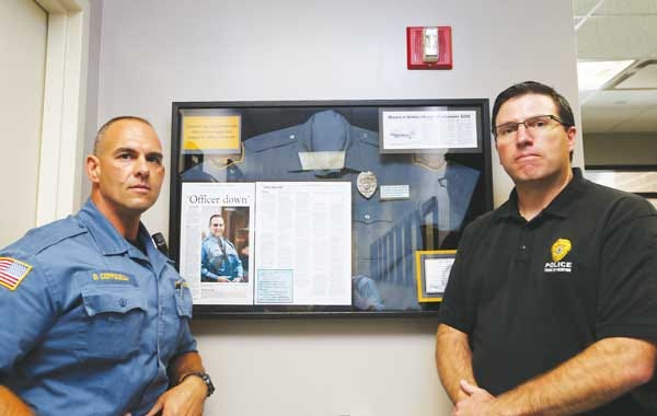 Officer Dean Coppolella, left, and Chief Michael Richards pose in front of a glass display case at Newton Police Department on Tuesday. The display contains mementos and news clippings from an unsolved hit and run Coppolella was a victim of in Aug. 2005.