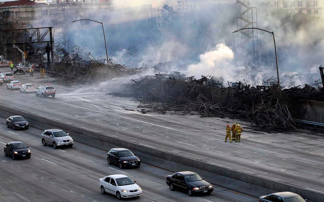 Los Angeles firefighters and state highway workers watch as an apartment building under construction smolders after a massive fire along State Route 110 that engulfed the site near downtown Los Angeles on Monday, Dec. 8, 2014. Crews battled two large fires in Los Angeles early Monday, including one downtown that closed portions of two major freeways and blanketed the area in ash and heavy smoke. (AP Photo/Nick Ut)