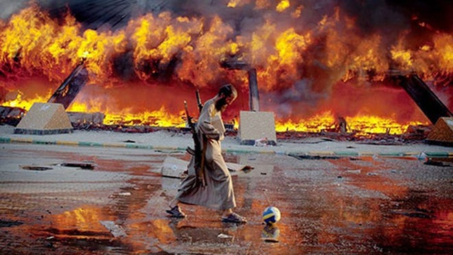 A photo from the ‘Seeing Double’ exhibit shows an armed rebel fighter kicking a soccer ball near a compound.