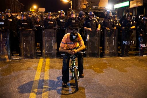 A man sits on a bicycle in front of a line of police officers in riot gear ahead of a 10 p.m. curfew in the wake of Monday's riots following the funeral for Freddie Gray, Tuesday, April 28, 2015, in Baltimore. (AP Photo/David Goldman)