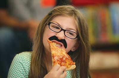 Photo by Daniel Freel/New Jersey Herald - Andrina Ismail, wearing her pizza man moustache, enjoys the best part of the day as she takes a big bite of her pizza slice.
