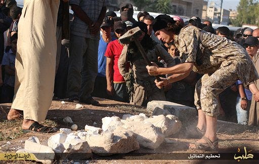 In this image posted on a militant website by the Aleppo branch of the Islamic State group on Friday, July 3, 2015, a militant is seen smashings items that the group claims are smuggled archaeological pieces from the historic central town of Palmyra, Syria. An IS statement says the busts were found when the smuggler was stopped at a checkpoint. The case was later referred to an Islamic court, which ordered that the pieces be destroyed and the man be whipped. (militant website via AP)