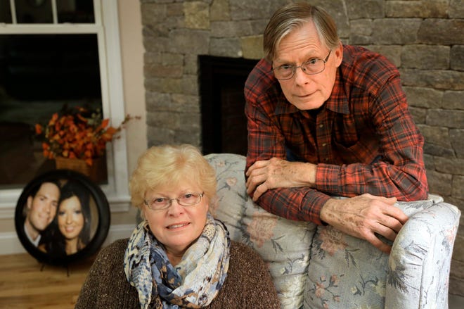 In this Monday, Nov. 9, 2015 photo, Rosanne Carruthers, center, and her husband Robert Carruthers, of Sterling, Mass., pose at their home in Sterling near a photo of their son Neil Carruthers and his wife, Tina. The young couple died within two days of each other, and it took more than a year for the medical examiner's office to determine Neil's cause of death. A chronic shortage of forensic pathologists across the country has created a backlog of hundreds of cases, causing long delays in autopsy results and the issuance of death certificates. (AP Photo/Steven Senne)