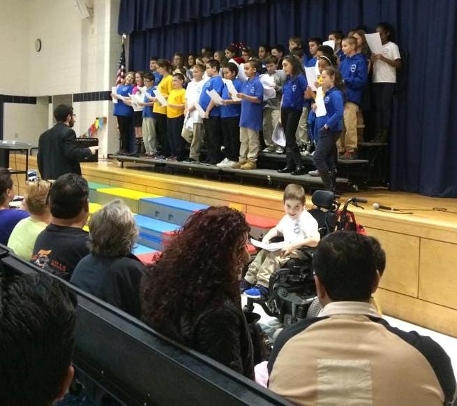 This image, a public post from Cidalia Maria's Facebook page, shows Camron Silva, in wheelchair, sitting off the stage nearby his classmates at Spencer Borden.