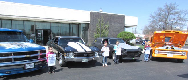 Vintage vehicles like the ones here, lined up at the State Bank of Toulon Kewanee Branch, will be parked on the downtown Kewanee streets on July 16 for the car show that will be held during the Prairie Chicken Festival. The children are holding posters advertising a 50-50 drawing for which tickets are being sold now.