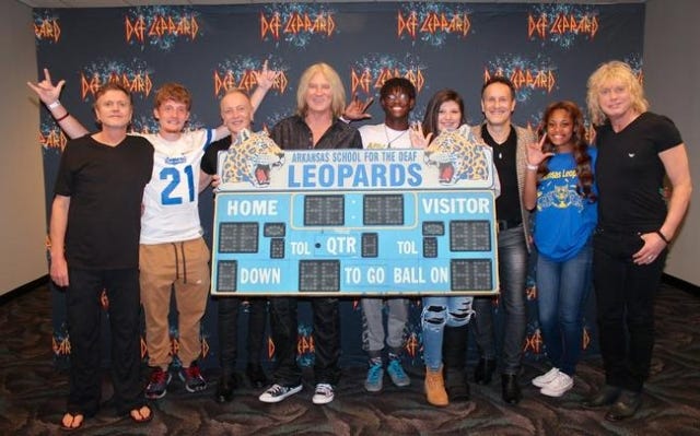 Members of the rock band Def Leppard pose Wednesday with students from the Arkansas School for the Blind with a reproduction of the school‘s scoreboard, which reads, "Arkansas School for the Blind Deaf Leopards," in Verizon Arena in North Little Rock, where Def Leppard was playing a concert. Photo courtesy of Def Leppard