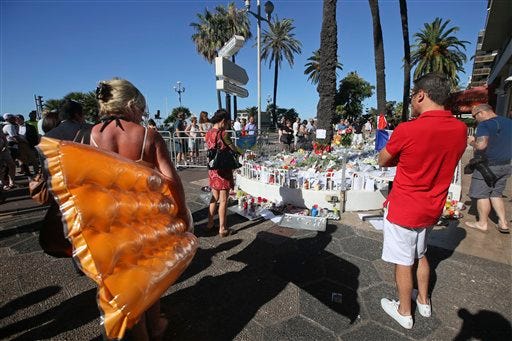 Tourists and citizens stand by flowers laid near the scene of a truck attack in Nice, southern France, Saturday, July 16, 2016. Nice's seaside boulevard reopens to traffic Saturday following a dramatic truck attack which killed more than 80 people and wounded more than 200 others at a fireworks display. (AP Photo/Luca Bruno)