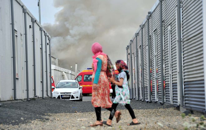 Refugees walk past containers as smoke billows in background in the refugee camp near Kassel, Germany, on Sunday. German authorities say 16 asylum seekers suffered from smoke inhalation after a fire broke out at a container housing facility in the central city of Kassel. The Kassel fire department told the dpa news agency the fire started inside one of the residences Sunday and that an anti-migrant attack had been ruled out.