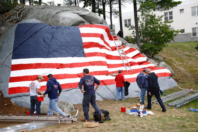 Off-duty Weymouth firefighters repaint a large rock covered in a US flag at Weymouth High. The stars will be painted after the blue field is dry.