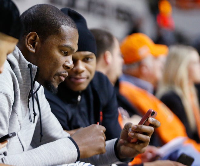 Oklahoma City Thunder's Kevin Durant, left, and teammate Russell Westbrook, right, look at Durant's phone during a break in the action as they watch an NCAA college basketball game between Kansas and Oklahoma State in Stillwater, Okla., Saturday, March 1, 2014. (AP Photo/Sue Ogrocki)