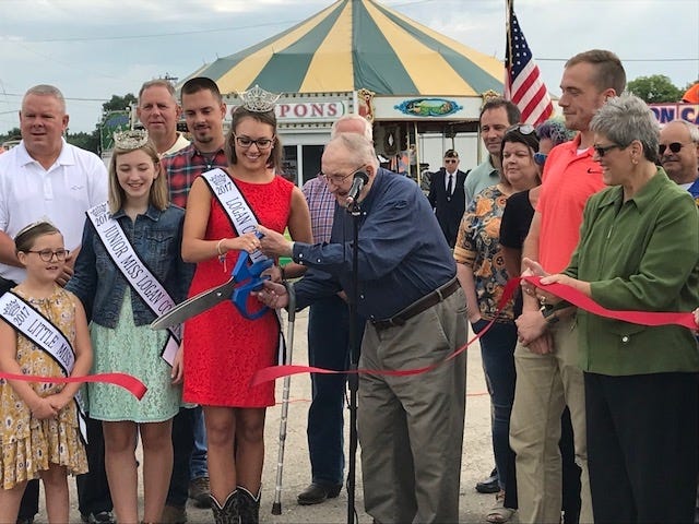 Logan County Fair welcomed visitors Tuesday morning with a ribbon cutting ceremony near the front gates of the fairgrounds. The fair which runs until Sunday, Aug. 5 is one of the biggest events held in Logan County. Logan County Fair Association board president Dean Bruns, on the right holding the scissors, opens the 2018 fair along with Queen Meg Meeker, left, had the honor of cutting the ribbon while other city and county dignitaries witnessed the ceremony. [Photo by Jean Ann Miller/The Courier]