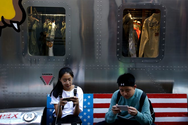 Shoppers sit on a bench with a decorated with U.S. flag browsing their smartphones outside a fashion boutique selling U.S. brand clothing at the capital city's popular shopping mall in Beijing, Monday, Sept. 24, 2018. China raised tariffs Monday on thousands of U.S. goods in an escalation of its fight with President Donald Trump over technology policy and accused Washington of bullying Beijing and damaging the global economy. (AP Photo/Andy Wong)