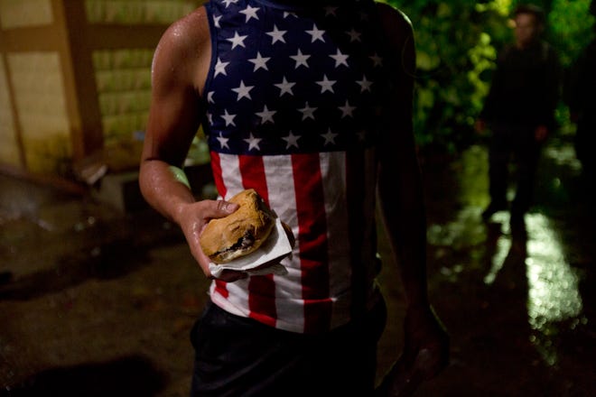 A Honduran migrant wearing a U.S. flag motif shirt, holds a sandwich at an improvised shelter in Chiquimula, Guatemala, Tuesday, Oct. 16, 2018. U.S. President Donald Trump threatened on Tuesday to cut aid to Honduras if it doesn't stop the impromptu caravan of migrants, but it remains unclear if governments in the region can summon the political will to physically halt the determined border-crossers. (AP Photo/Moises Castillo)