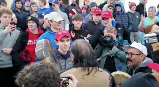 In this Friday image made from video provided by the Survival Media Agency, a teenager wearing a "Make America Great Again" hat, center left, stands in front of an elderly Native American singing and playing a drum in Washington. The Roman Catholic Diocese of Covington in Kentucky is looking into this and other videos that show youths, possibly from the diocese's all-male Covington Catholic High School, mocking Native Americans at a rally in Washington.