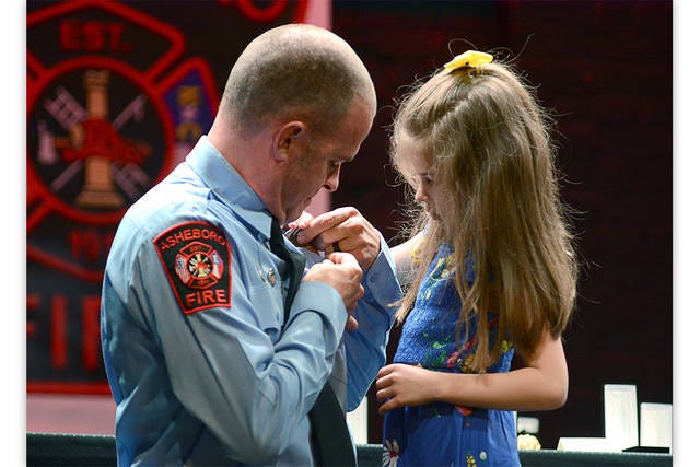 BADGE OF HONOR — Benjamin Hicks’ daughter, Harper, pins on his badge following his promotion to fire lieutenant during a ceremony recognizing Asheboro Fire Department members at the Sunset Theatre Thursday night. (Scott Pelkey / Special to The Courier-Tribune)