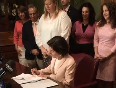 Gov. Gina Raimondo signs the abortion-rights bill into law Wednesday night following its passage by the Senate and then House. To her right is Sen. Erin Lynch Prata, who played a central role in guiding the measure through a contentious process. [The Providence Journal, from video / Katherine Gregg]